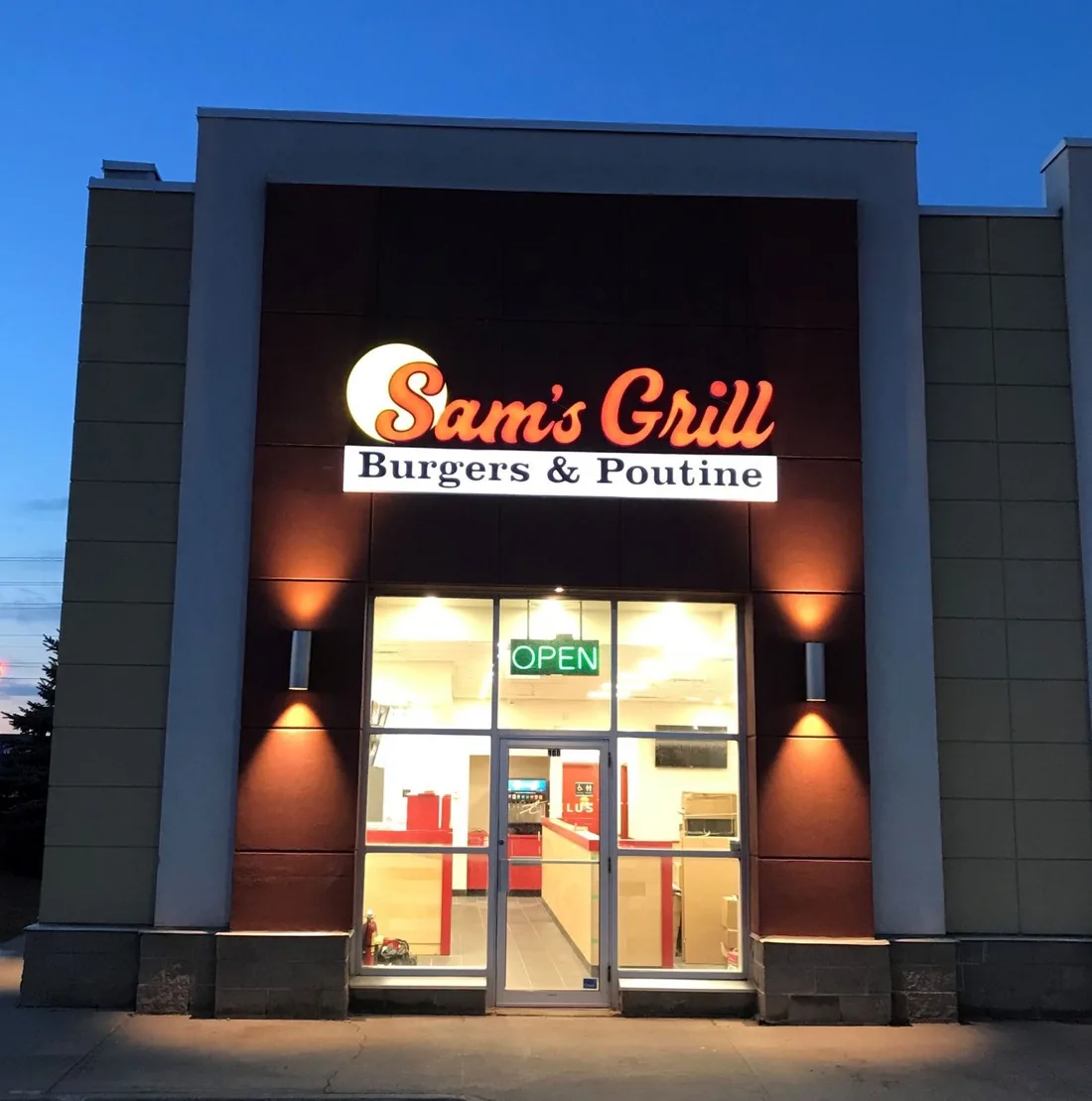 A Sam's Grill storefront at dusk, the open sign lit and the counter visible through the glass